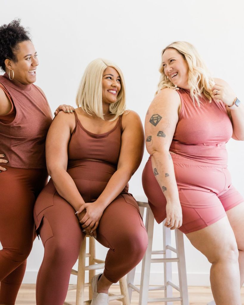 Three women dressed in matching brown athletic wear, sitting together and smiling against a light backdrop. The scene conveys a sense of community and body positivity.